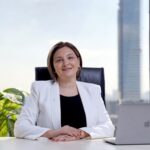 Professional woman smiling at desk with skyscraper view, representing corporate leadership.